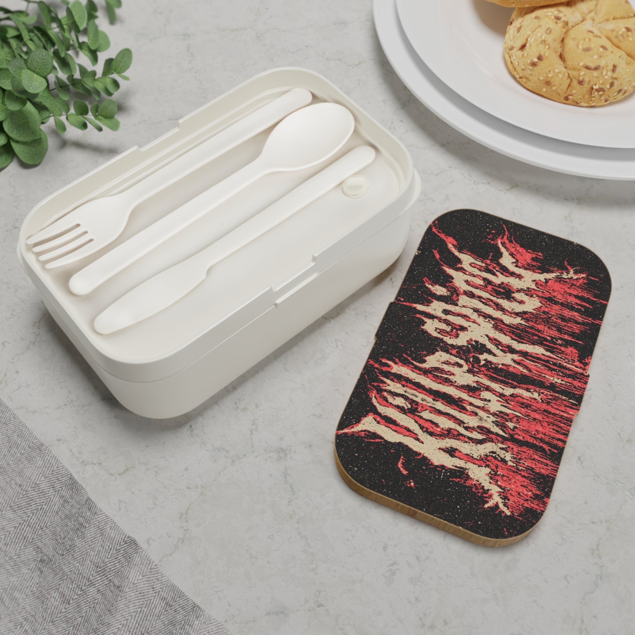 White cutlery set in a box on a gray surface with a decorative coaster featuring red and beige patterns.