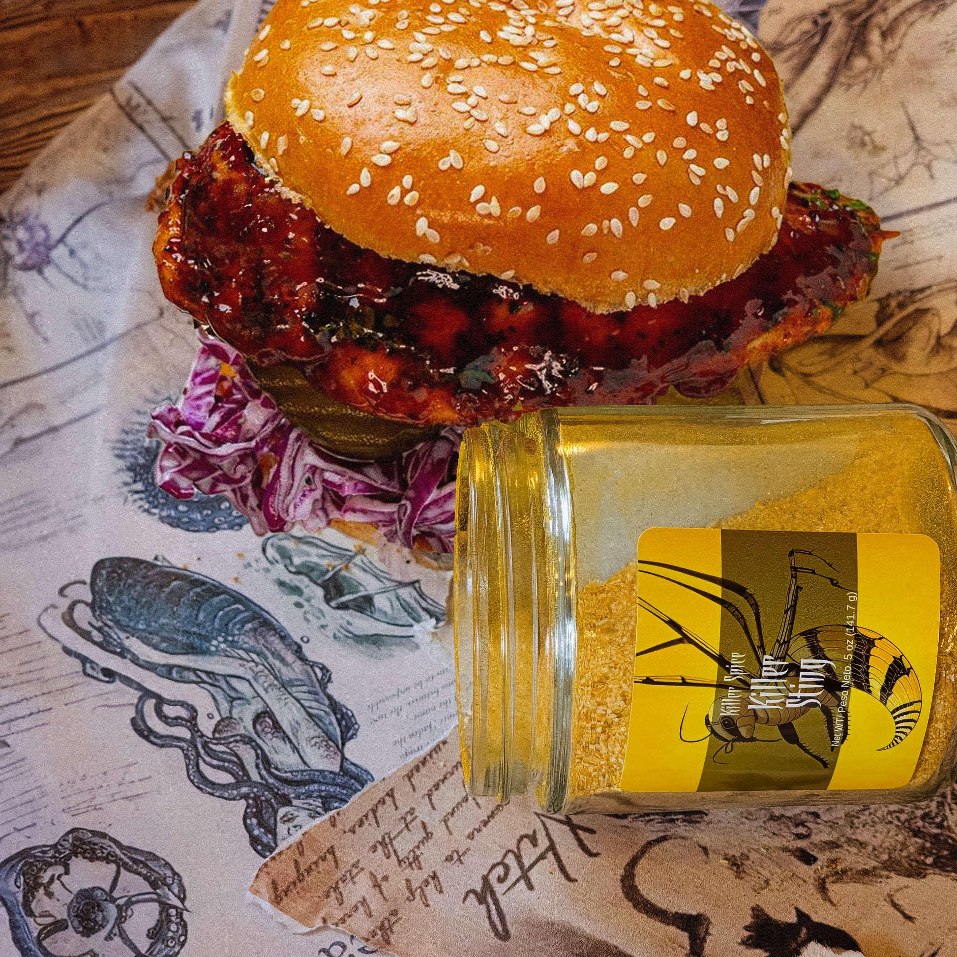A jar of Killer Sting honey from Killer Spice positioned next to a chicken sandwich with coleslaw and cayenne pepper on a table with patterned tablecloth.