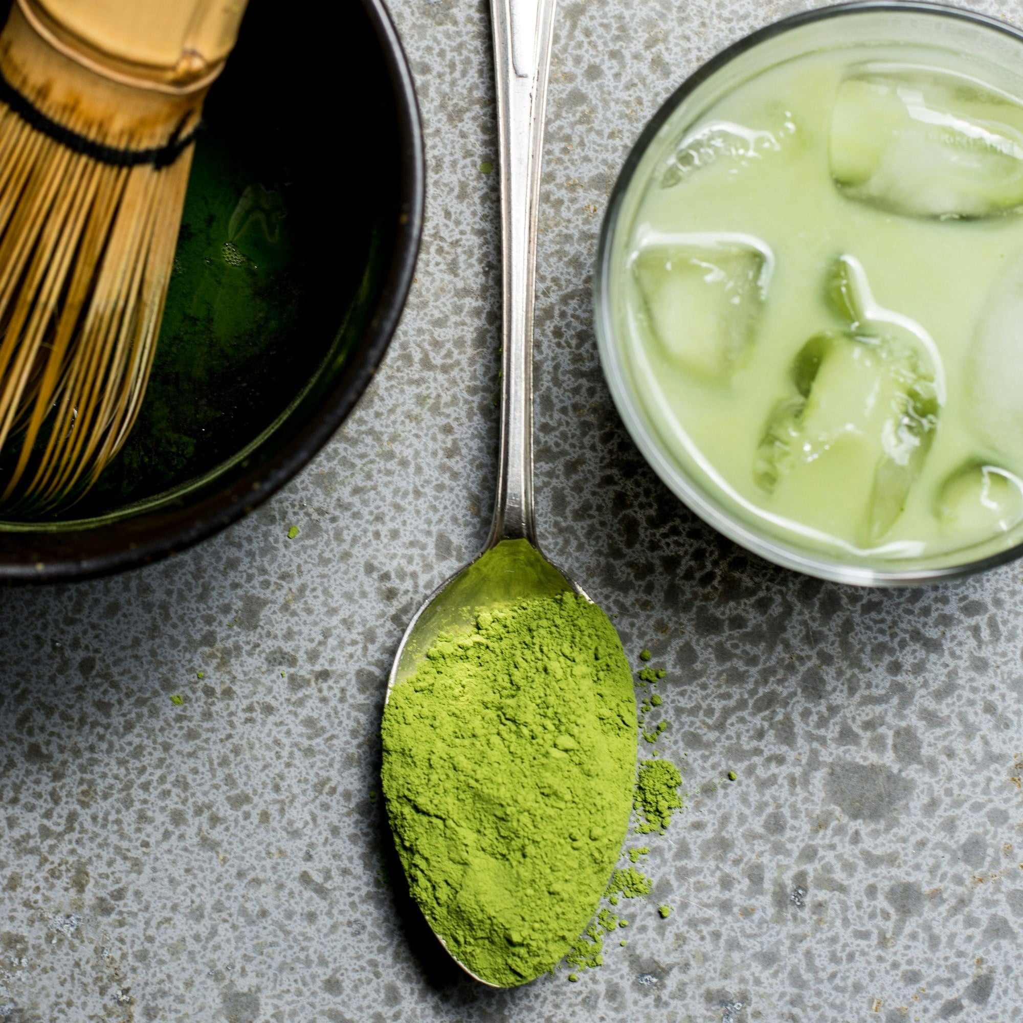 Spoon with green matcha powder, whisk, and glass of iced matcha on a gray surface