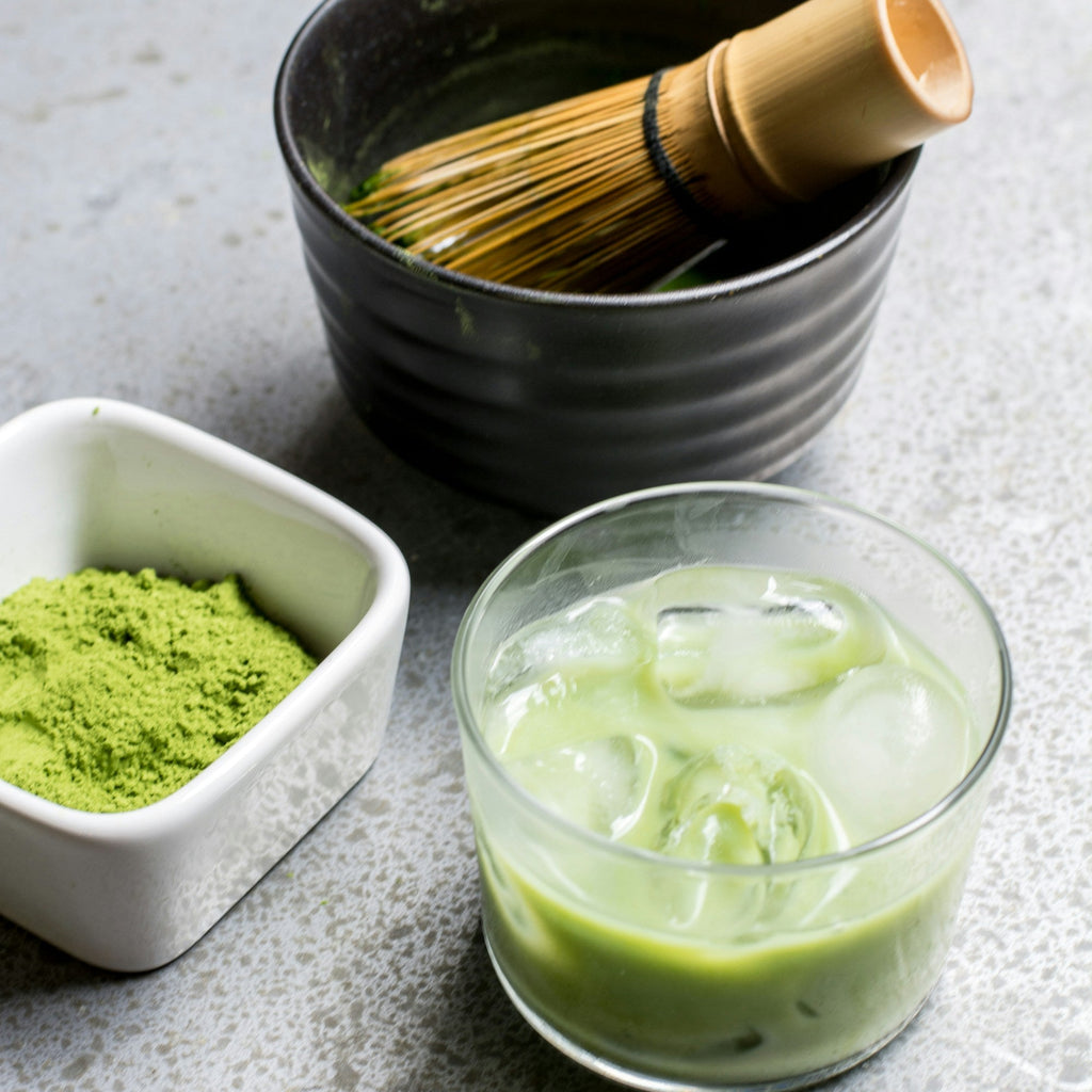 Matcha green tea preparation with matcha powder, whisk, and glass of iced matcha on a gray surface.