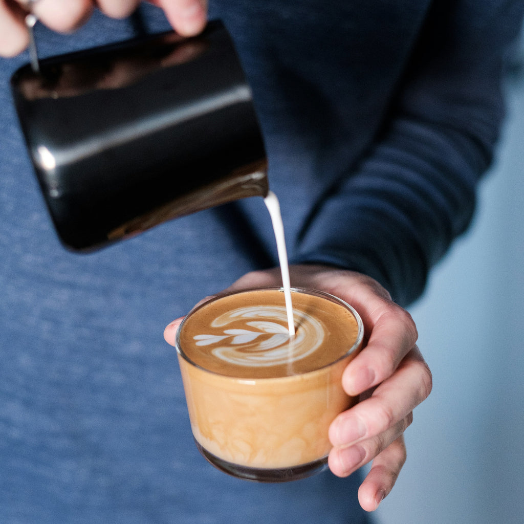 Person pouring milk into a coffee cup with latte art against a blue background