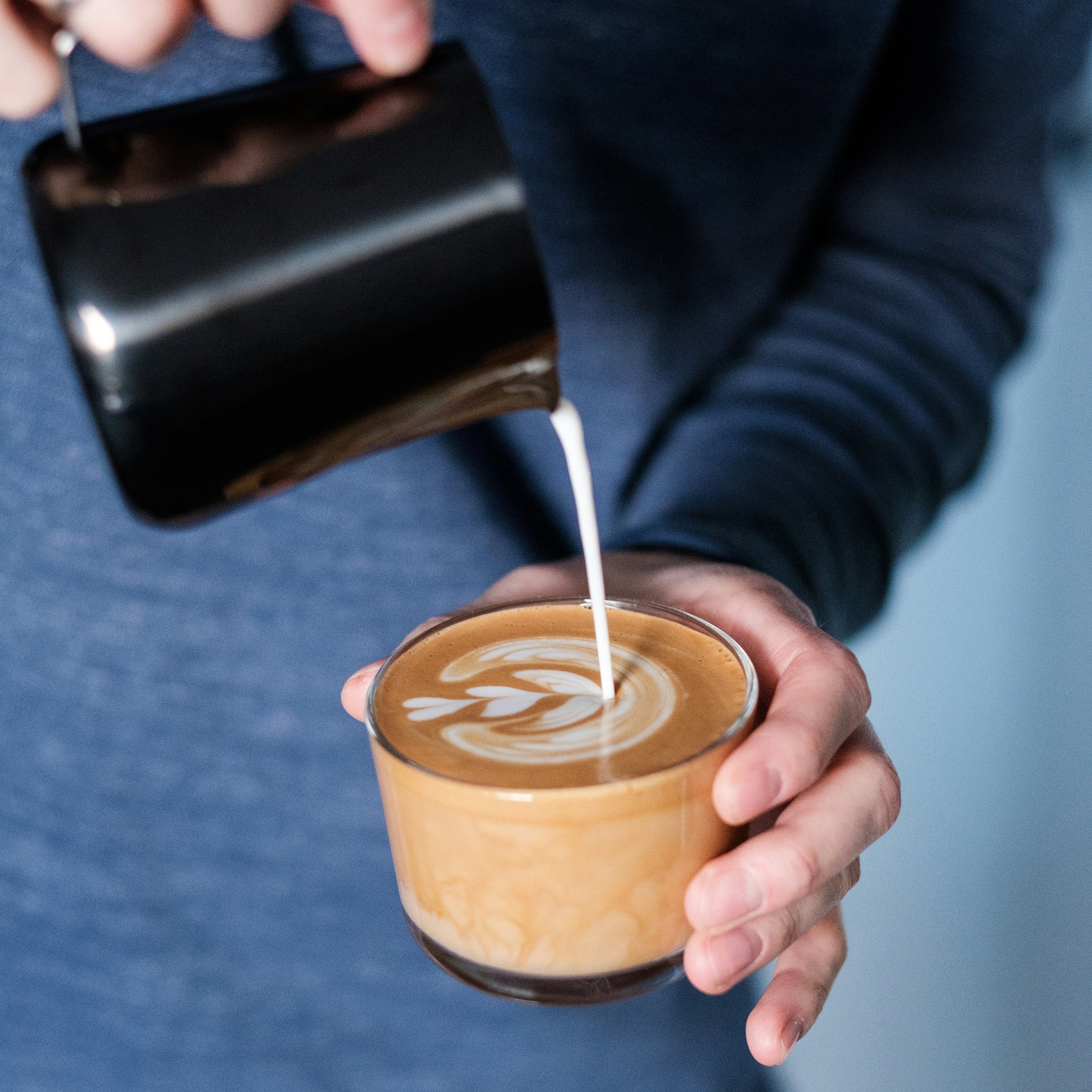 Person pouring milk into a coffee cup with latte art against a blue background