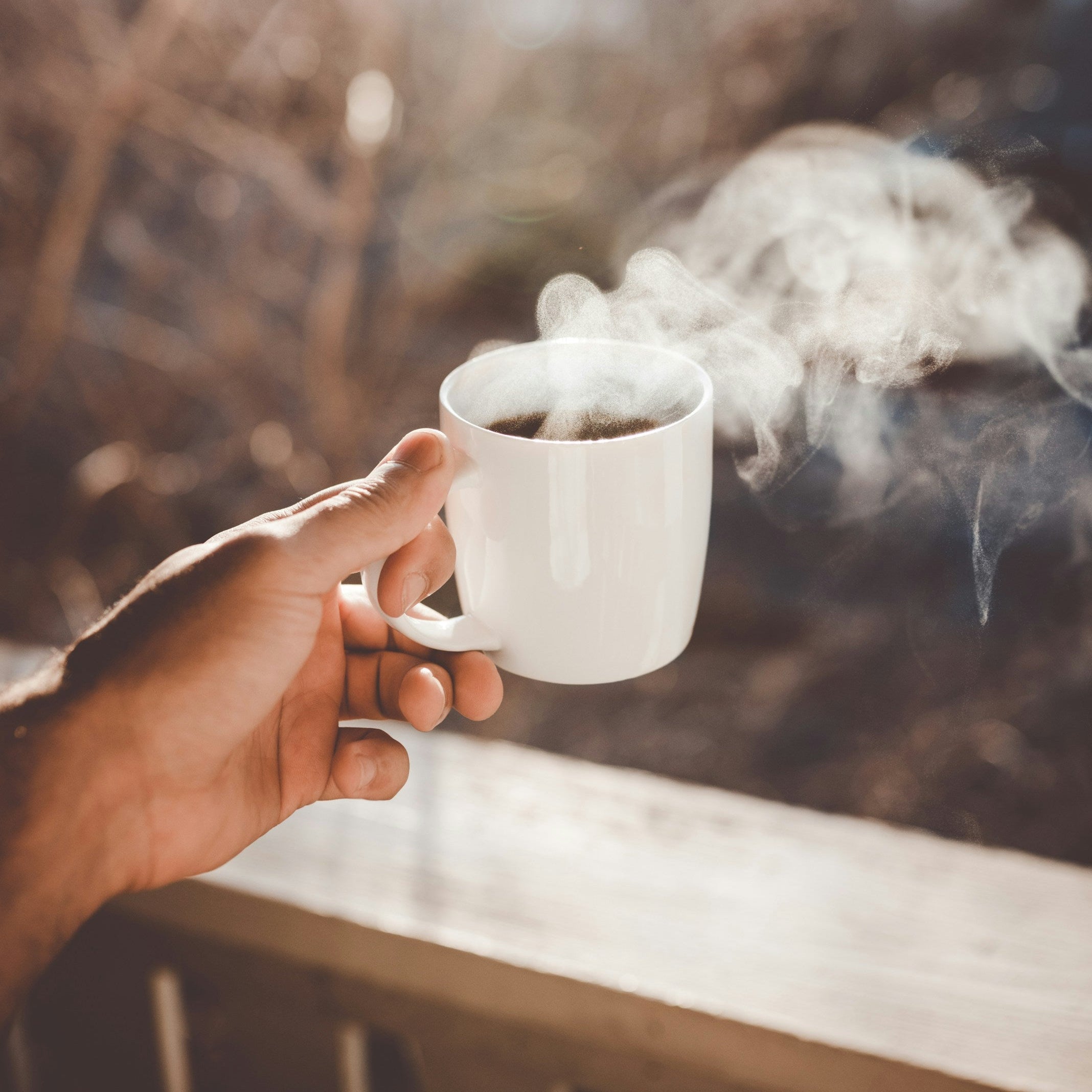 Hand holding a steaming white mug with a blurred natural background