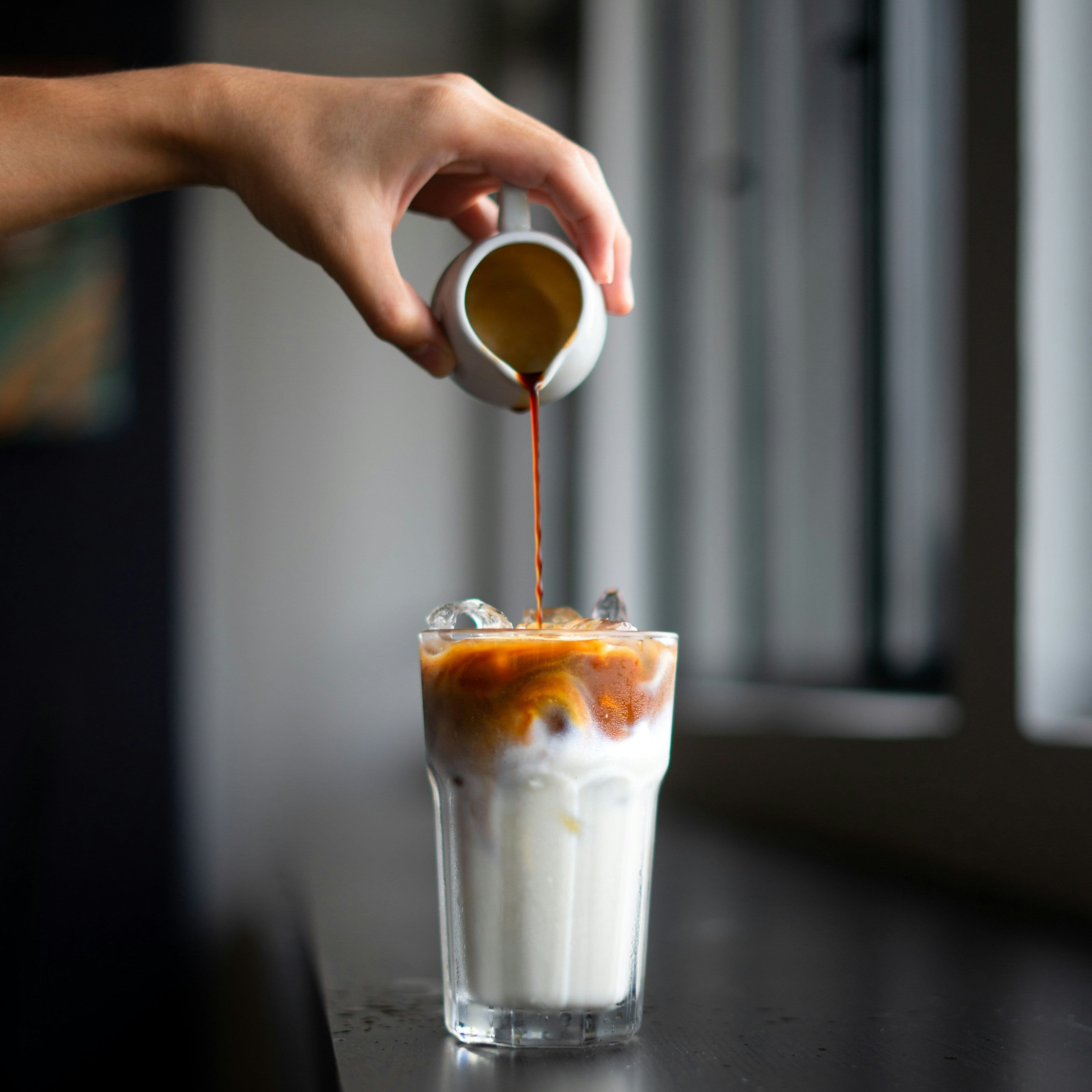 Hand pouring a creamy liquid into a glass of iced coffee on a dark surface.