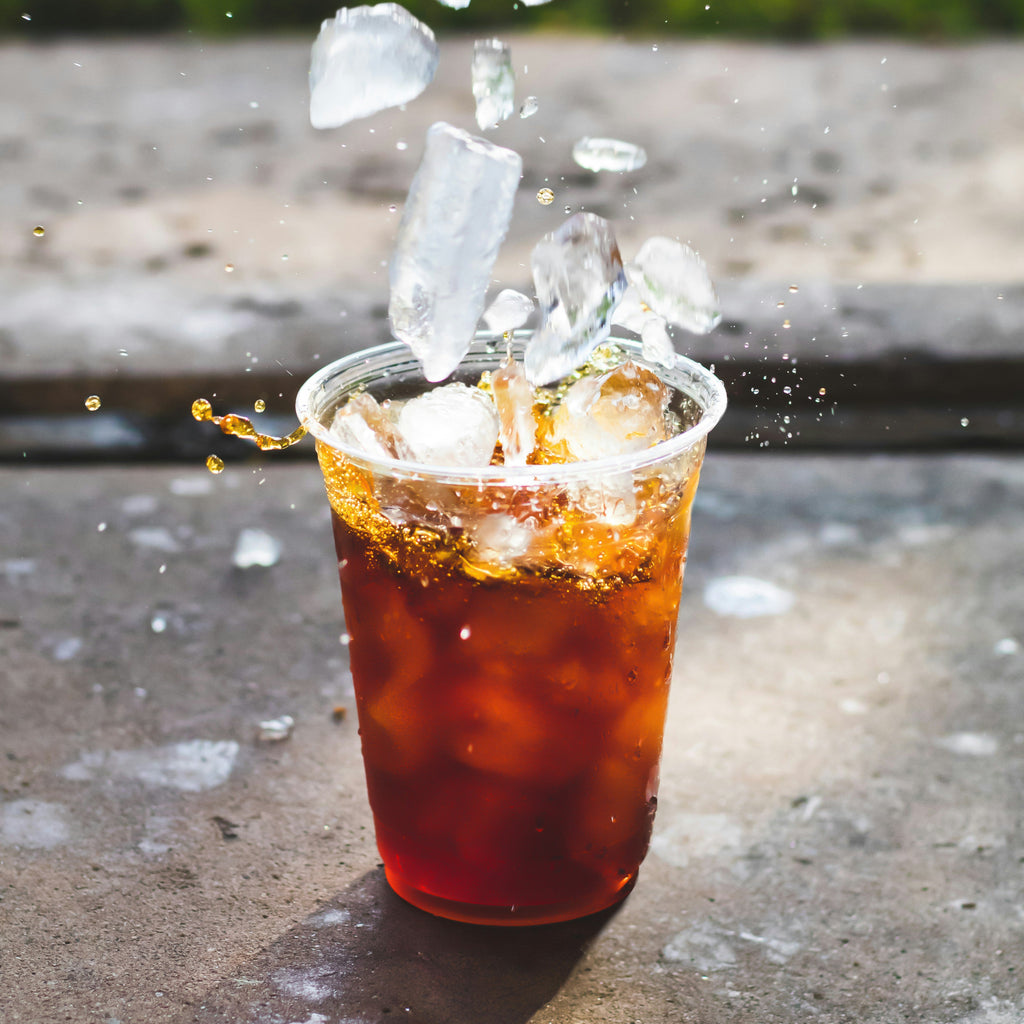 Glass of iced tea with ice cubes being added on a concrete surface with grass in the background