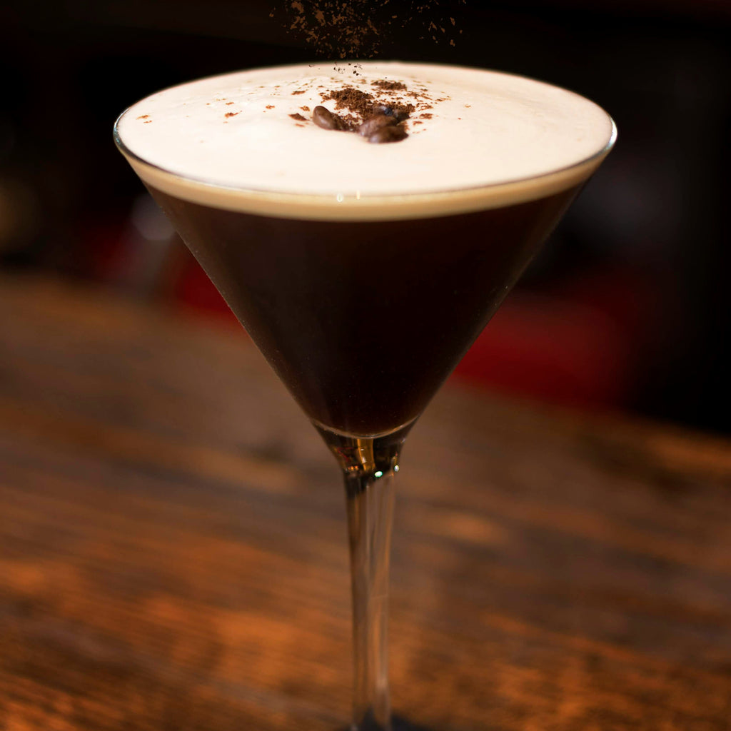 Dark cocktail with a white foam top in a martini glass on a wooden surface.