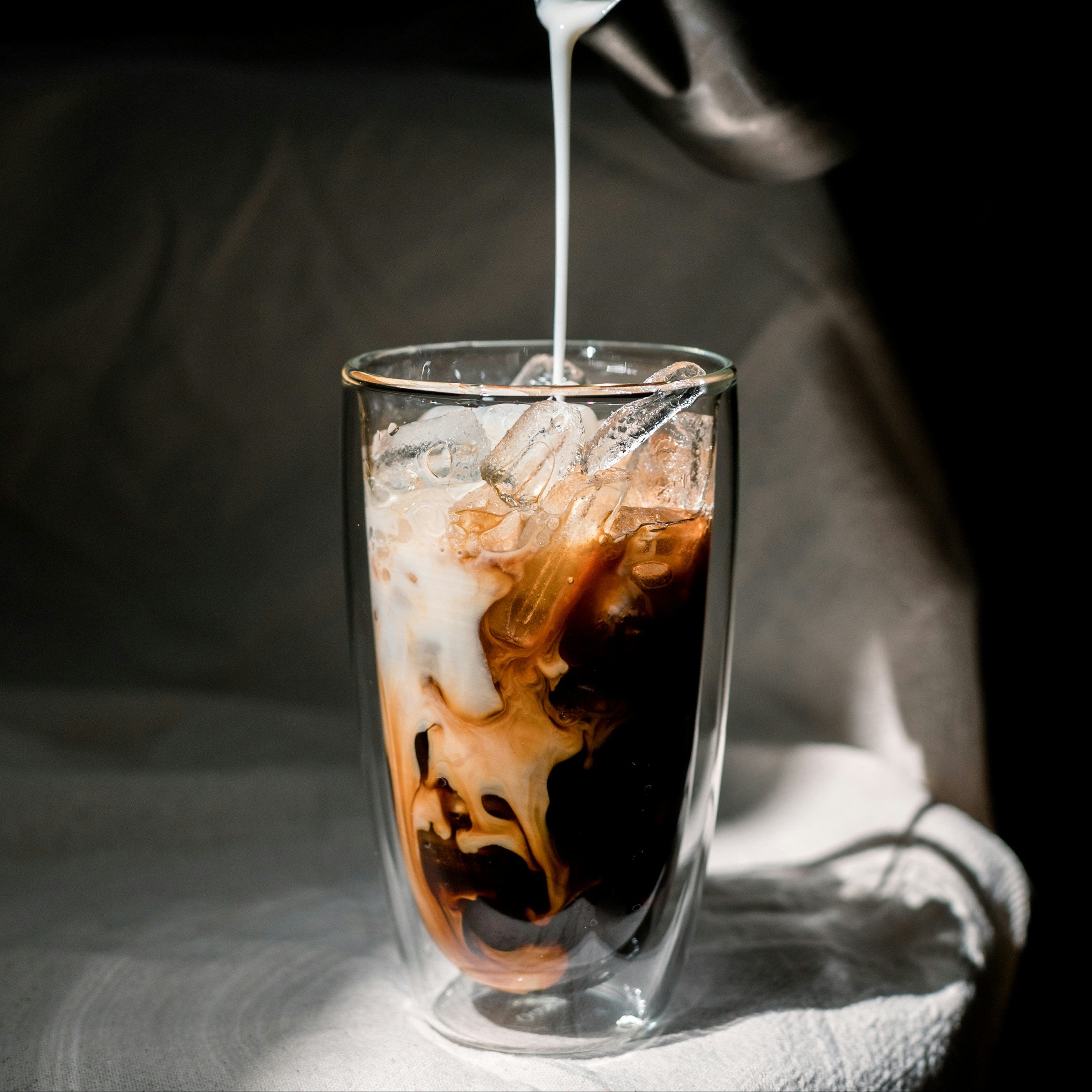 Glass of iced coffee with a spoon pouring cream against a dark background