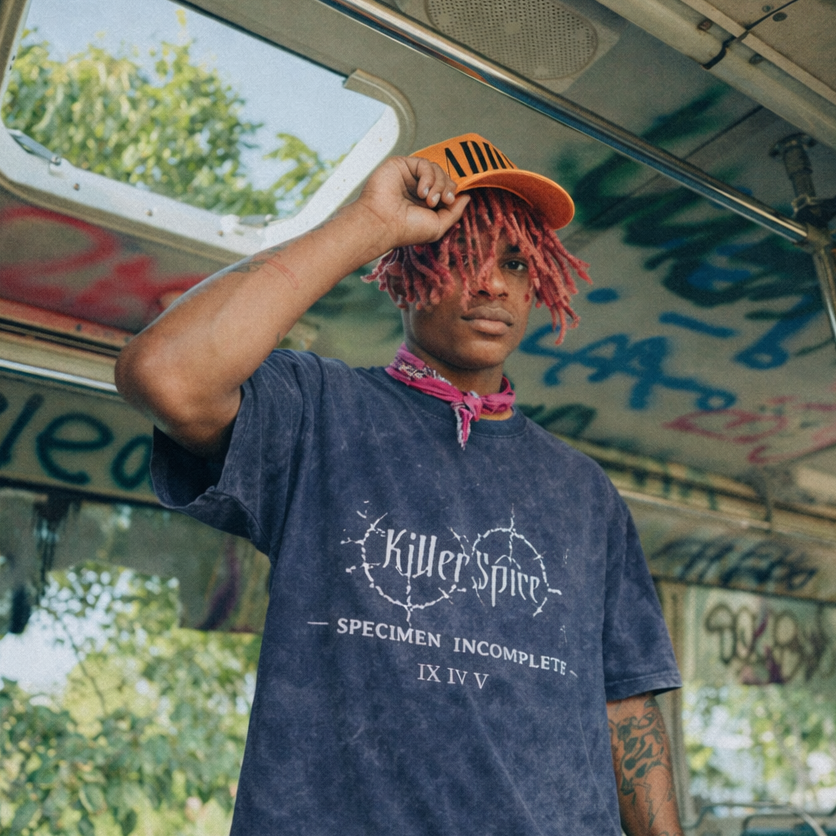 Person with pink hair wearing a blue t-shirt inside a graffiti-covered vehicle.