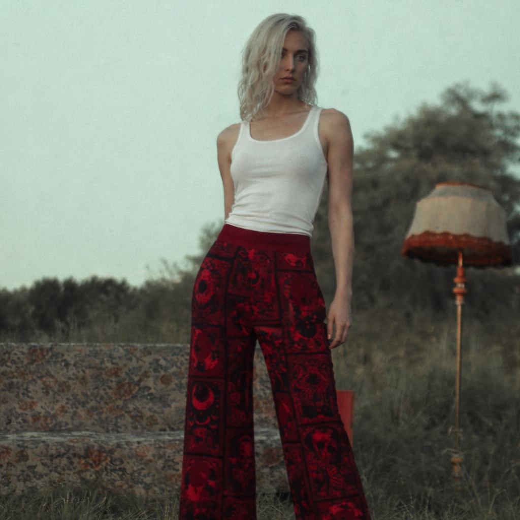 Woman wearing a white tank top and red patterned pants standing in a grassy field with a lamp in the background.