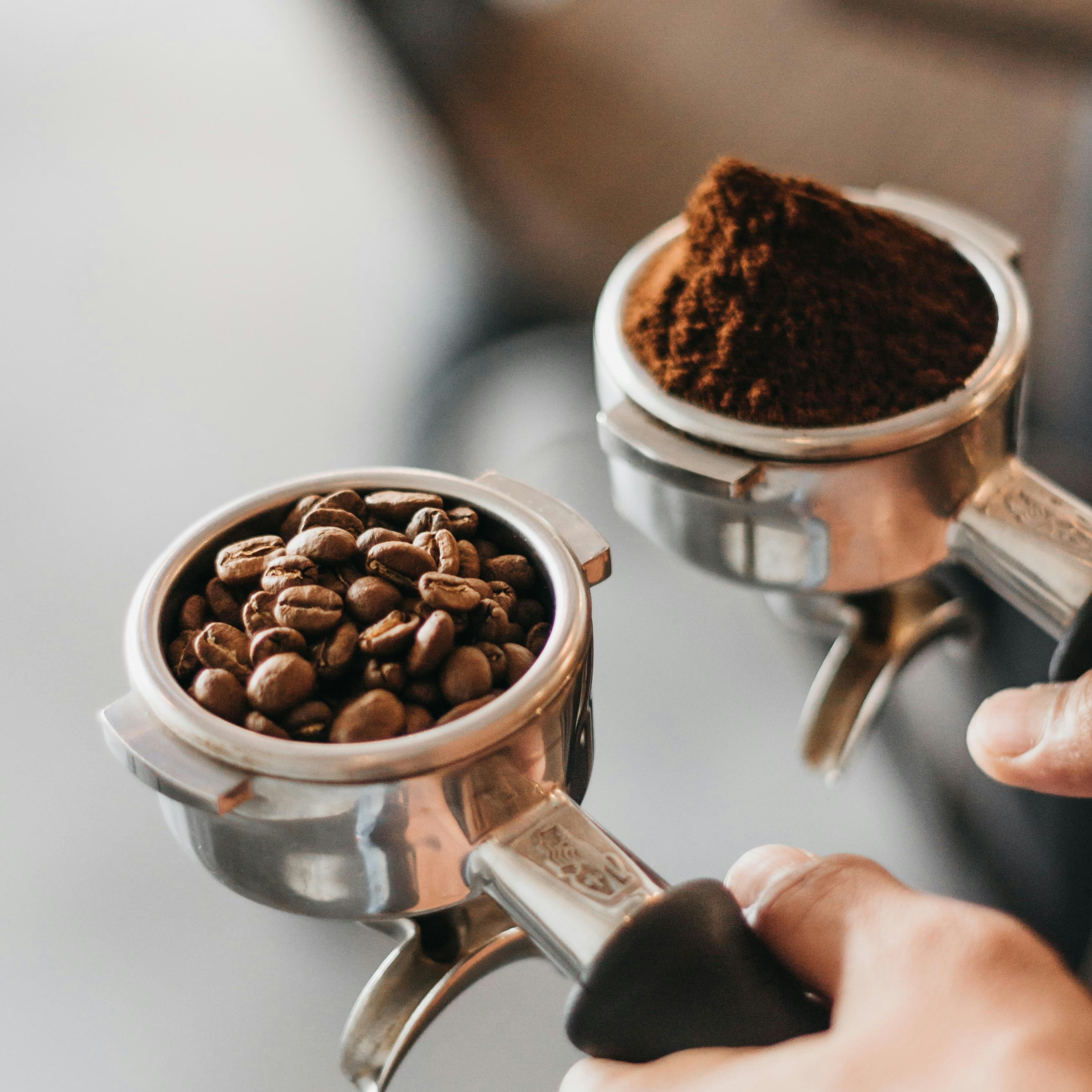 Two portafilters filled with coffee beans and ground coffee, held by a hand.